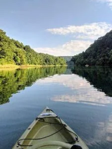 Kayak bow pointing down a calm river with tree-lined banks and clouds reflected on the water.