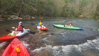 Four kayakers navigating a rocky river rapid in a wooded area.