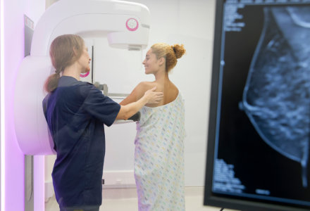 Healthcare professional positions a patient for a mammogram using a digital mammography machine in a clinical imaging room. The patient stands facing the machine wearing a patterned medical gown while the clinician adjusts the patient’s shoulder and upper body for proper alignment. A computer monitor on the right displays a grayscale breast imaging scan, and the room is brightly lit with clean, modern diagnostic equipment visible.