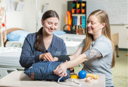 Two people work at a table in a clinical classroom setting. One person tightens a blood‑pressure cuff on the other person’s forearm while additional therapy and healthcare tools—such as a squeeze ball, tape, and small assistive items—are arranged on the table. In the background, a hospital bed, linens, and medical training equipment are visible under bright classroom lighting.