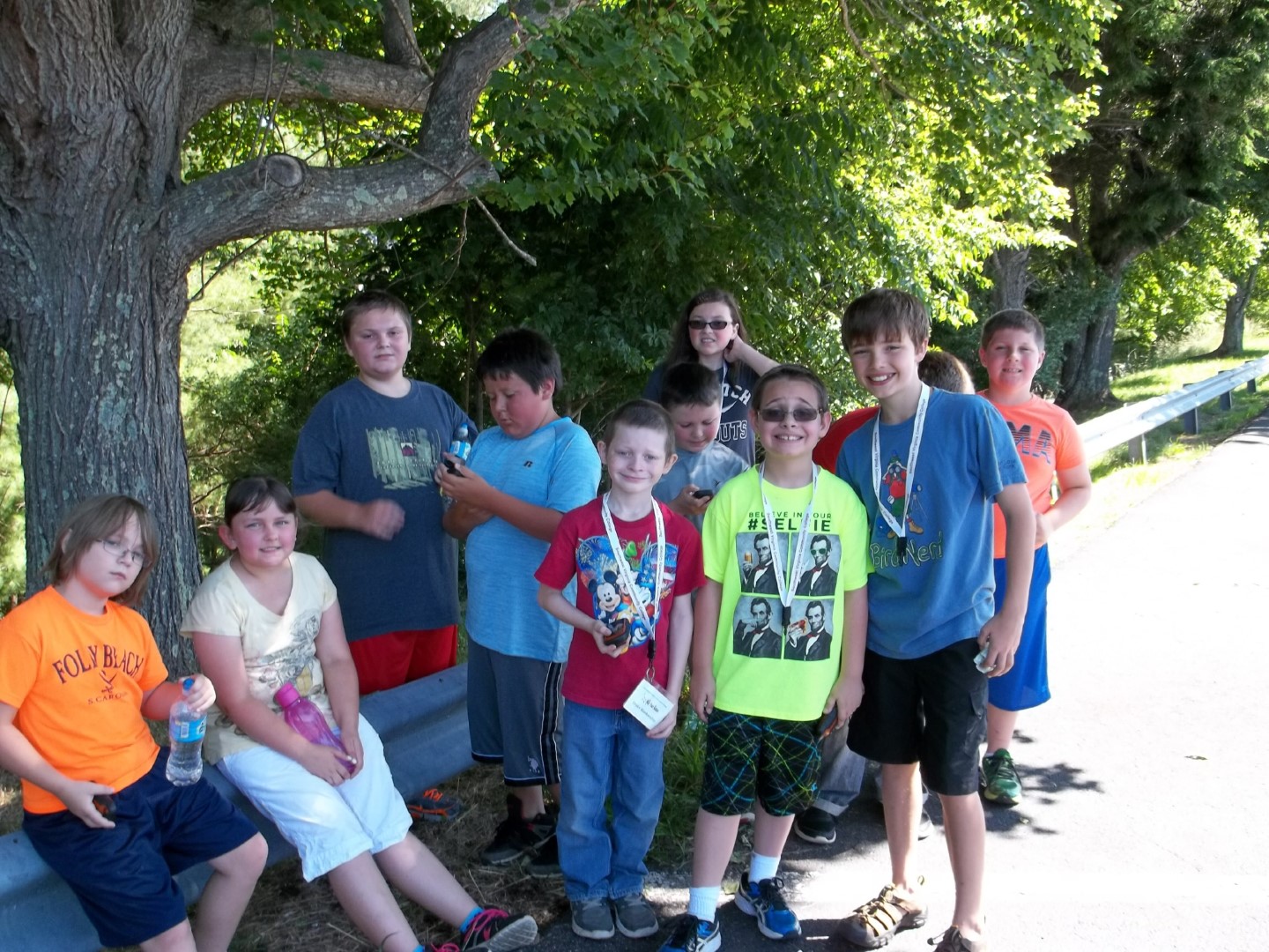 Group of kids standing and sitting by a roadside guardrail under a large tree on a sunny day.
