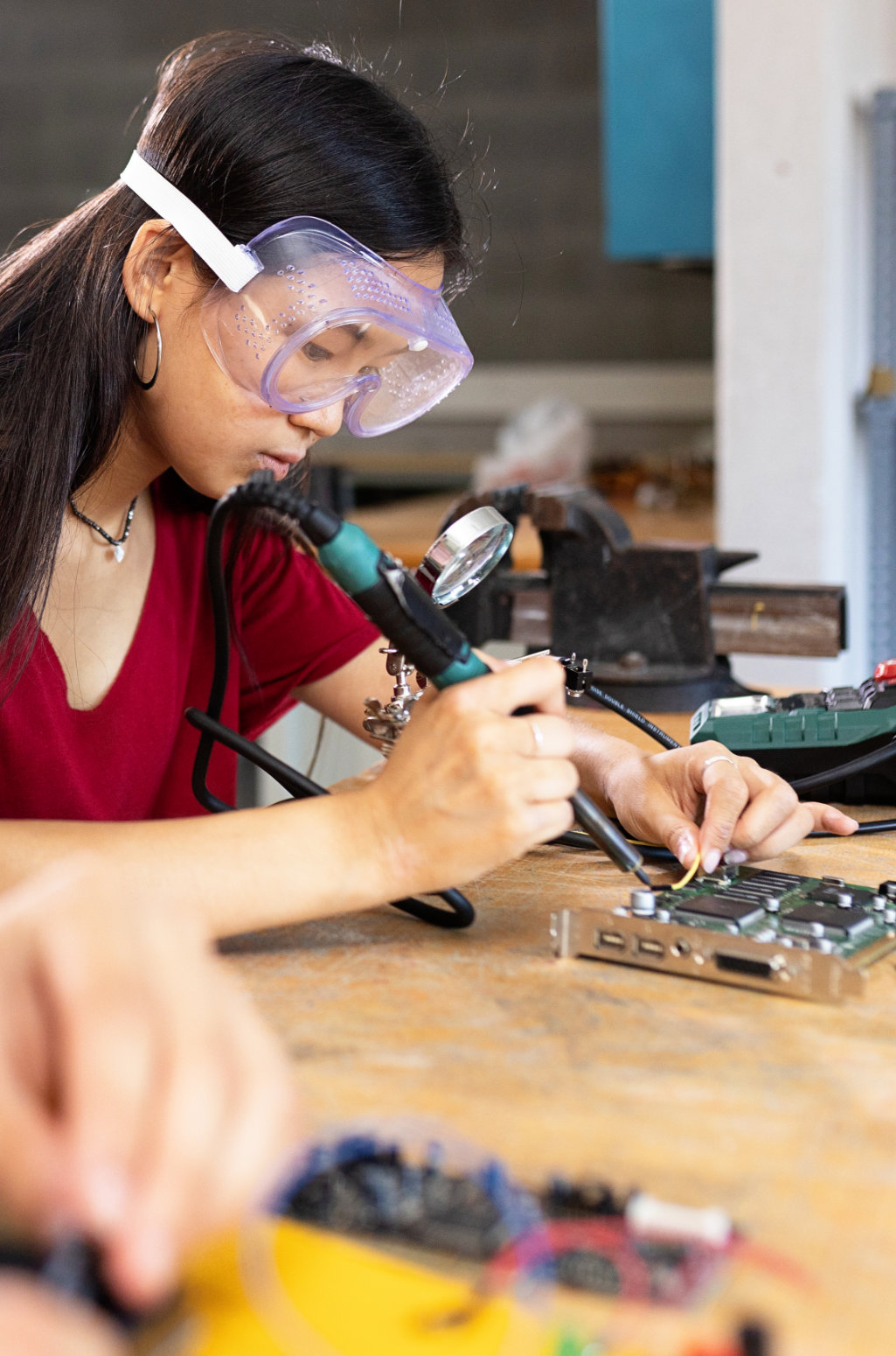 Student wearing clear safety goggles uses a soldering iron to work on a circuit board at a workshop table. The circuit board and electronic components rest on a wooden work surface, with tools, wires, and equipment visible in the foreground and background, suggesting a hands-on electronics or technology training environment.