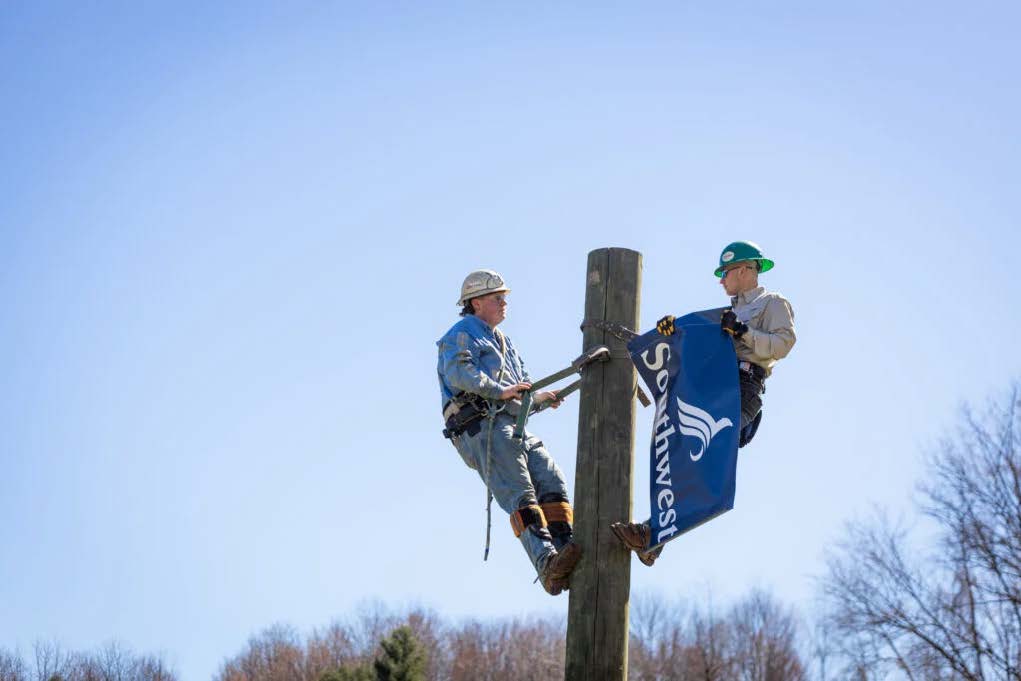 Two workers wearing safety gear are secured to a tall wooden utility pole while attaching a blue banner that reads “Southwest” against a clear sky.