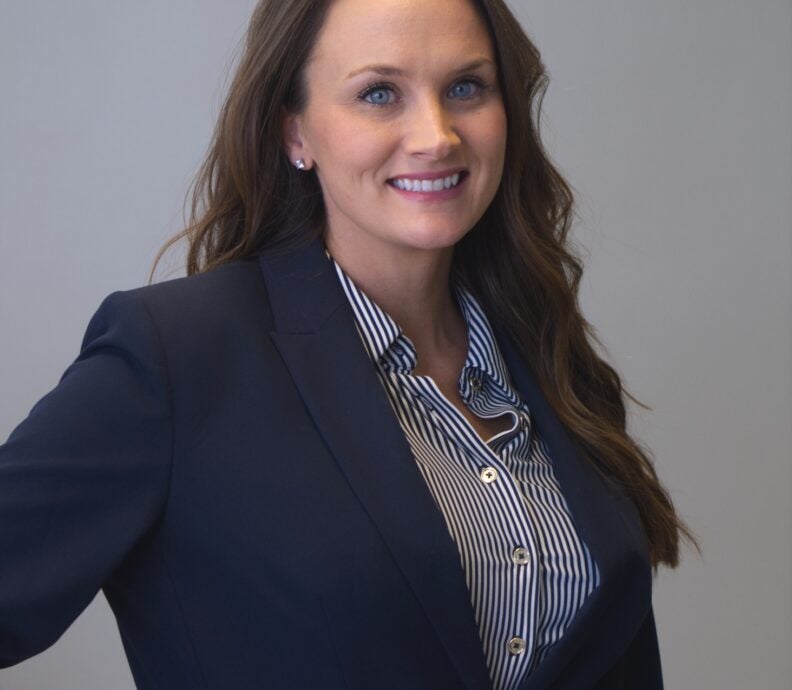 Dr. Bethany Rose wearing a dark navy business suit with a striped button‑down shirt standing against a plain, light-colored background. Dr. Rose's posture is confident, with one hand resting on the hip. The suit jacket features a single visible button and a notched lapel, and her long, wavy hair falls over one shoulder.