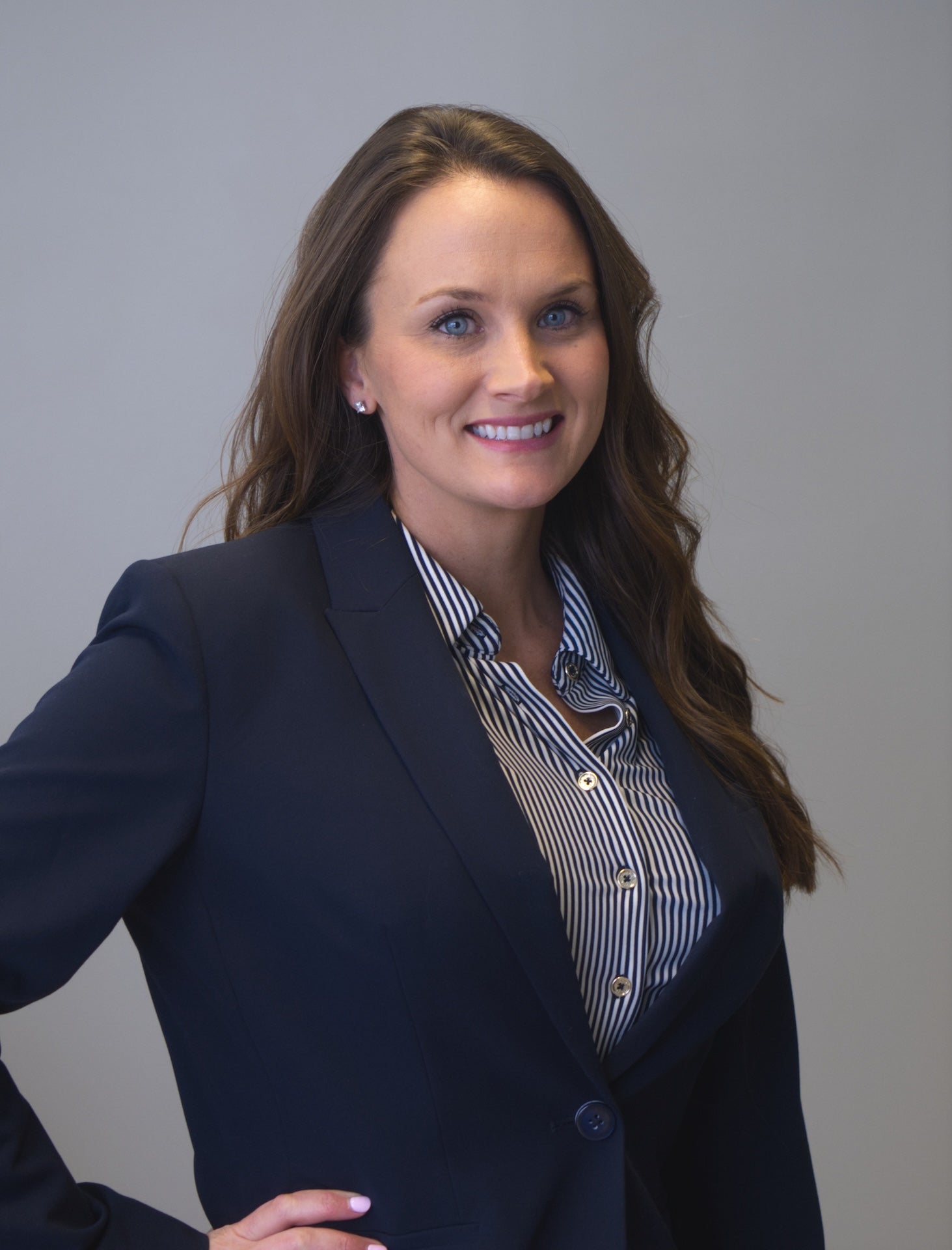 Dr. Bethany Rose wearing a dark navy business suit with a striped button‑down shirt standing against a plain, light-colored background. Dr. Rose's posture is confident, with one hand resting on the hip. The suit jacket features a single visible button and a notched lapel, and her long, wavy hair falls over one shoulder.